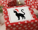 Black image of a silhouette of a cute cat wearing a bright red Santa hat with fluffy white trim and pom-pom detail. The cat stands alert with its tail curled and head turned to the side, giving it a poised, festive look.